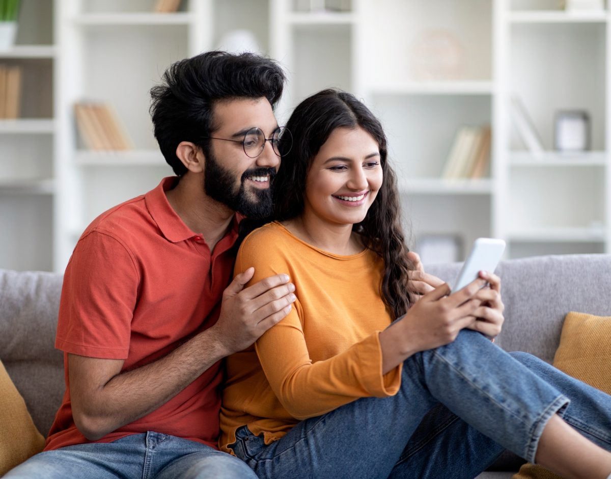 Smiling Young Indian Spouses Relaxing With Smartphone On Couch In Living Room