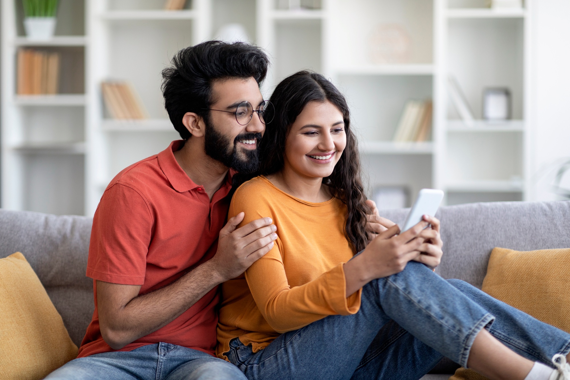 Smiling Young Indian Spouses Relaxing With Smartphone On Couch In Living Room